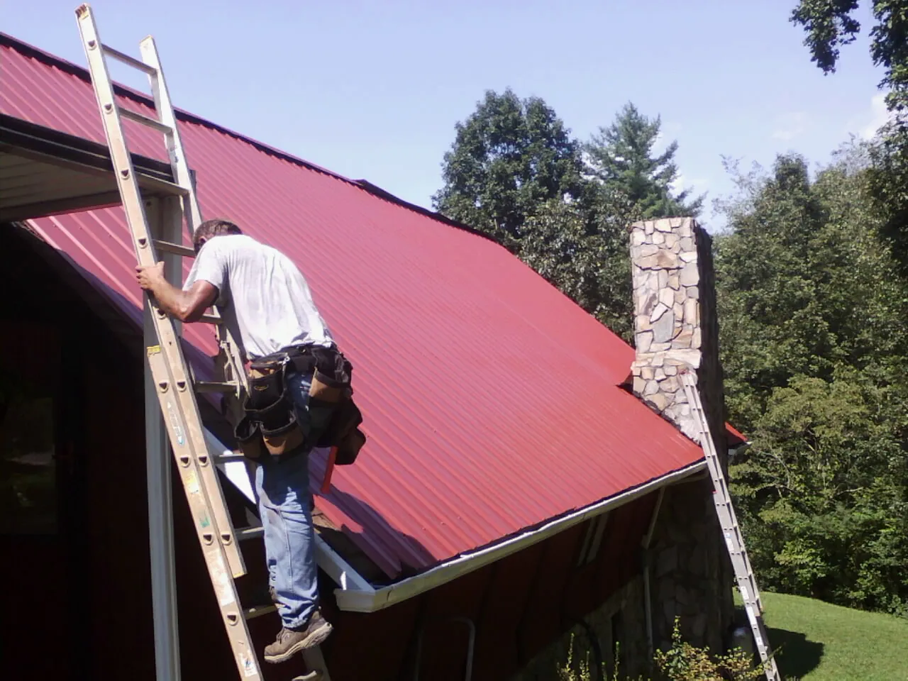 Roofer on a ladder beside a red metal roof and stone chimney — Soffit & Fascia Repair by Roofing Brothers in Loveville